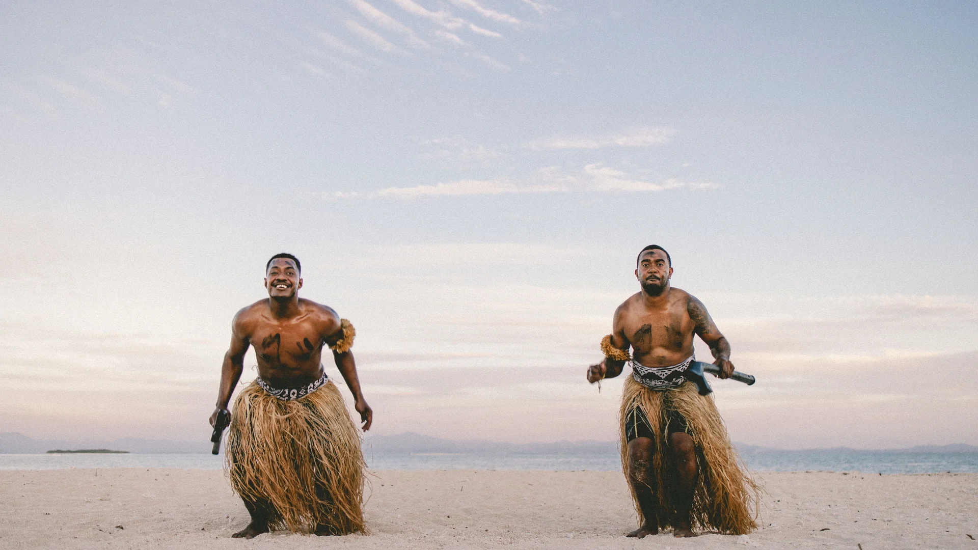 Two Fijian men in traditional warrior dress on a beach near Nadi, Fiji, at sunset