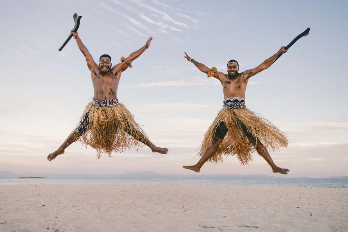 Two Fijian men in warrior dress leaping on a beach near Nadi, Fiji